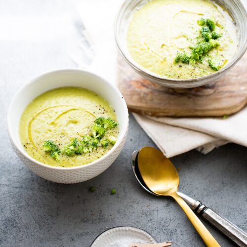 Dos tazones de sopa verde cremosa, probablemente crema de brócoli, con aceite de oliva y hierbas aromáticas. El tazón más pequeño reposa sobre una superficie gris junto a una cuchara de oro, un tenedor de plata y un salero. La escena es luminosa y acogedora.