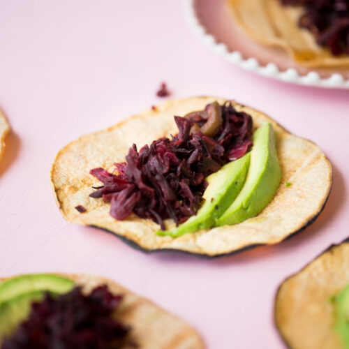 Una tortilla de maíz con col morada rallada y rebanadas de aguacate reposa sobre una superficie rosa claro. Al fondo se ven más tacos de flor de jamaica con ingredientes similares, uno de ellos en un plato blanco.