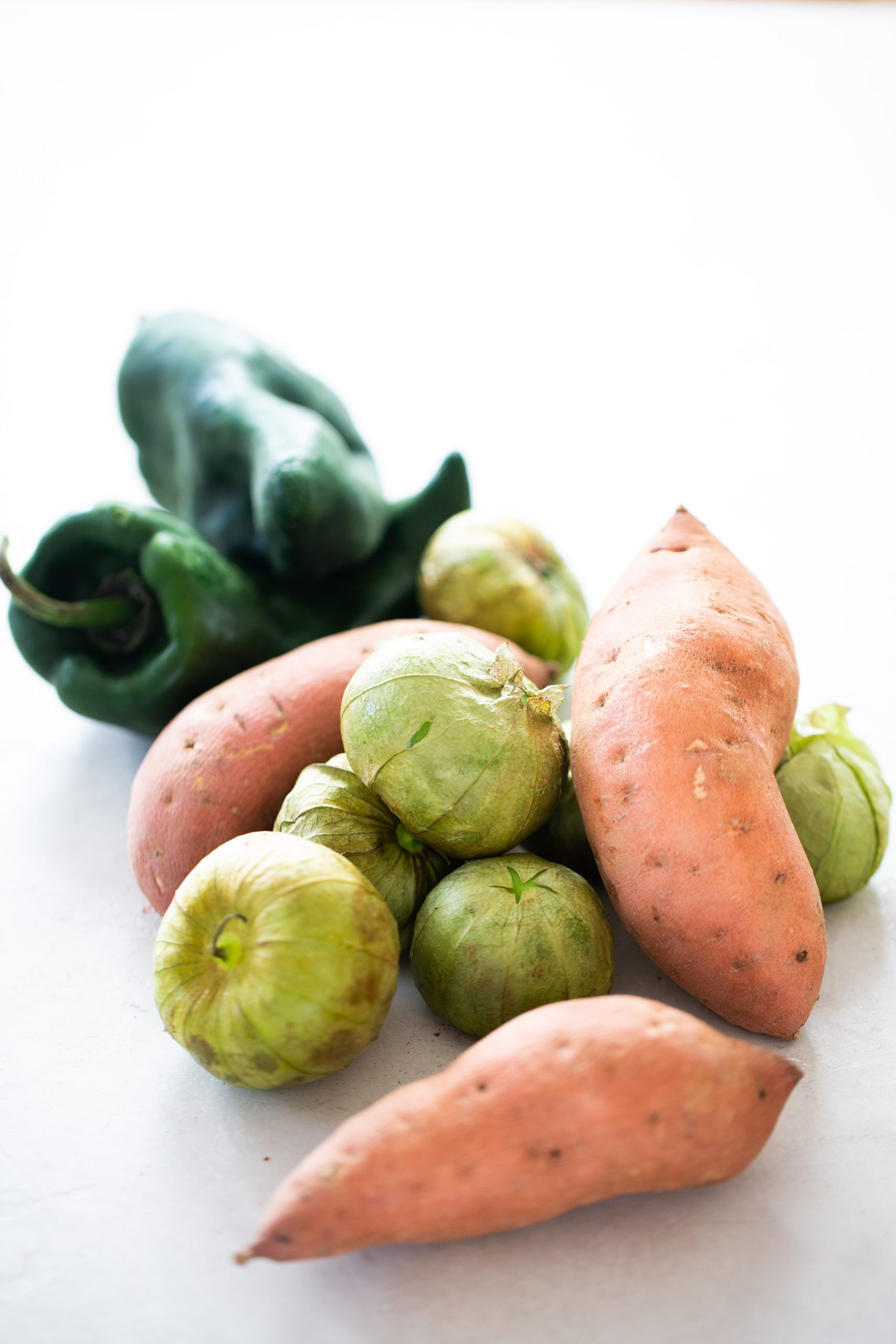 Tres camotes, varios tomatillos verdes con cáscara y dos chiles poblanos verde oscuro se disponen sobre una superficie blanca: ingredientes frescos perfectos para hacer enchiladas de camote, resaltando sus variadas texturas y formas naturales.
