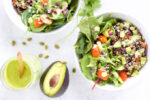 Dos tazones blancos rebosan de espinacas frescas, tomates cherry, aguacate en cubos, quinoa y col morada sobre una superficie blanca. Un aguacate partido por la mitad yace junto a un pequeño frasco de aderezo de cilantro y menta y una cuchara de madera, mientras que unas pepitas esparcidas completan la vibrante escena.