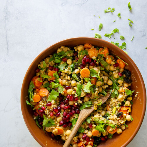 Una vista superior de un cuenco de cerámica marrón lleno de una ensalada colorida, que incluye verduras de hojas verdes picadas, zanahorias en rodajas, garbanzos y farro, semillas de granada y otras verduras mixtas. Una cuchara de madera descansa en el recipiente. Algunas hierbas picadas están esparcidas sobre la superficie de textura gris claro al lado del cuenco.