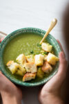 Una persona sosteniendo un plato de sopa de brócoli y espinaca verde adornado con picatostes.