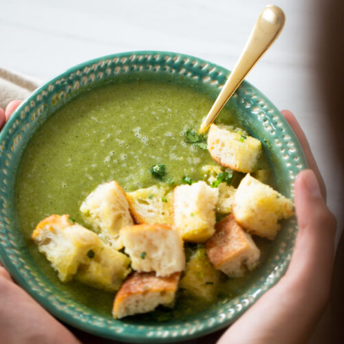 Una persona sosteniendo un plato de sopa de brócoli y espinaca verde adornado con picatostes.