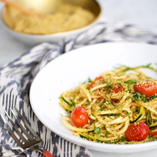 Un tazón blanco contiene espaguetis y pasta de calabacín con tomates cherry y hierbas verdes, rebozados en pan rallado dorado. El tazón reposa sobre un mantel estampado con un tenedor al lado. Al fondo, un pequeño tazón dorado contiene más pan rallado con una cuchara de madera.