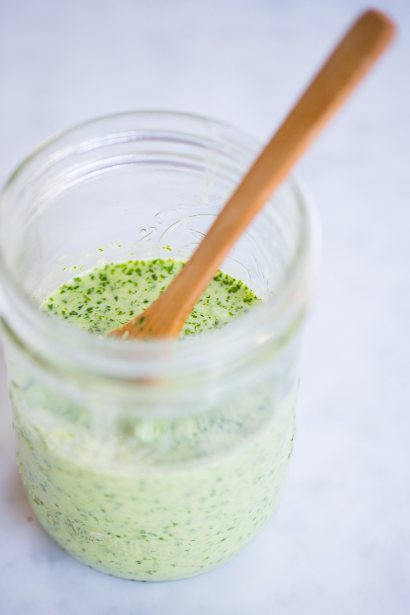 Un tarro de cristal lleno de aderezo de cilantro verde y cremoso sobre un fondo claro y borroso. Una cuchara de madera descansa dentro del tarro, parcialmente sumergida en el aderezo moteado. La mezcla parece espesa y casera.