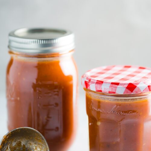 Dos frascos de vidrio llenos de salsa para enchiladas con chipotle se colocan uno al lado del otro sobre una superficie blanca. El frasco de la izquierda es más grande y está sellado con una tapa de metal, mientras que el frasco más pequeño de la derecha está cubierto con una tapa a cuadros rojos y blancos. Al frente, una cuchara de metal con residuos de salsa reposa sobre la superficie.