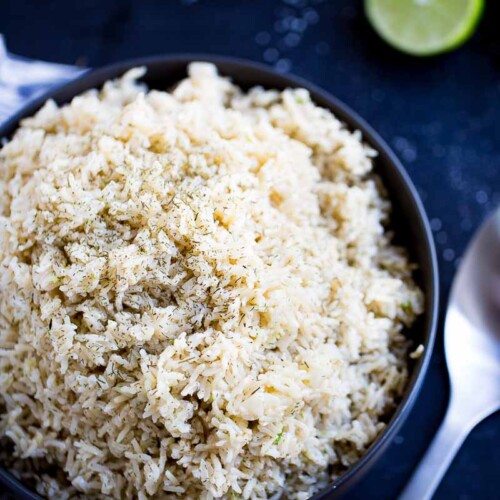 Un tazón negro lleno de esponjoso arroz integral, adornado con hierbas, reposa sobre una superficie oscura. Una cuchara de metal yace junto al tazón, y al fondo se ve una lima partida por la mitad, cerca de granos de arroz dispersos.