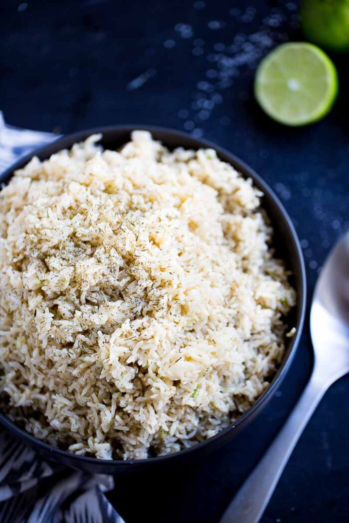 Un tazón negro lleno de esponjoso arroz integral, adornado con hierbas, reposa sobre una superficie oscura. Una cuchara de metal yace junto al tazón, y al fondo se ve una lima partida por la mitad, cerca de granos de arroz dispersos.