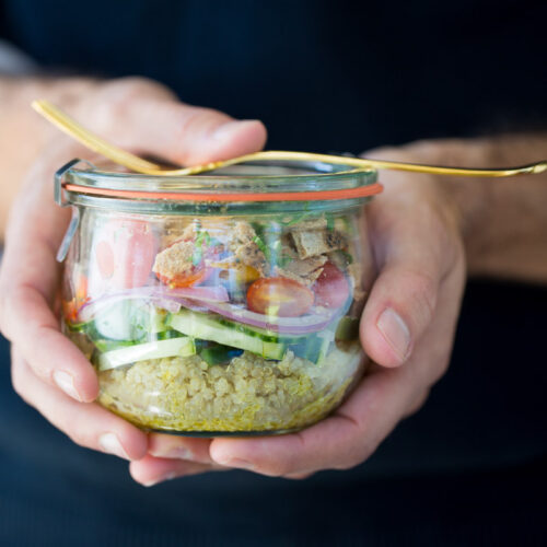 Una persona sostiene un frasco de vidrio lleno de una vibrante ensalada mediterránea de quinoa. Incluye capas de quinoa, pepinos en rodajas, tomates cherry y pollo a la parrilla. Un tenedor dorado está escondido debajo de la tapa. El fondo está difuminado, lo que enfatiza el colorido contenido de esta delicia saludable.