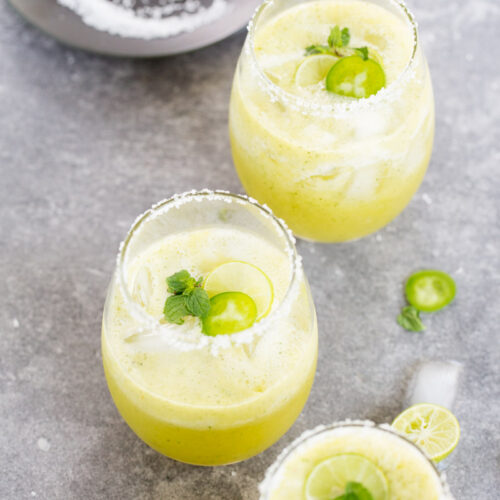 Tres vasos de margaritas frías de color amarillo verdoso, adornados con rodajas de lima, rodajas de jalapeño y menta fresca. Los bordes están cubiertos de sal. Un plato negro al fondo contiene una lima y sal. Rodajas circulares de lima y jalapeño se encuentran esparcidas sobre la superficie gris, enmarcando a la perfección la escena de la margarita.