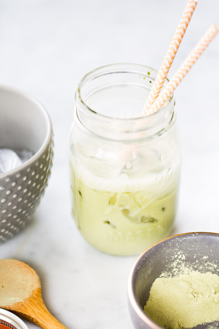 Un tarro lleno de matcha latte helado descansa sobre una superficie blanca, con dos pajitas estampadas asomando. Cerca, un bol gris con lunares rebosa de hielo, junto a una cuchara de madera y un pequeño cuenco de polvo de matcha verde vibrante.
