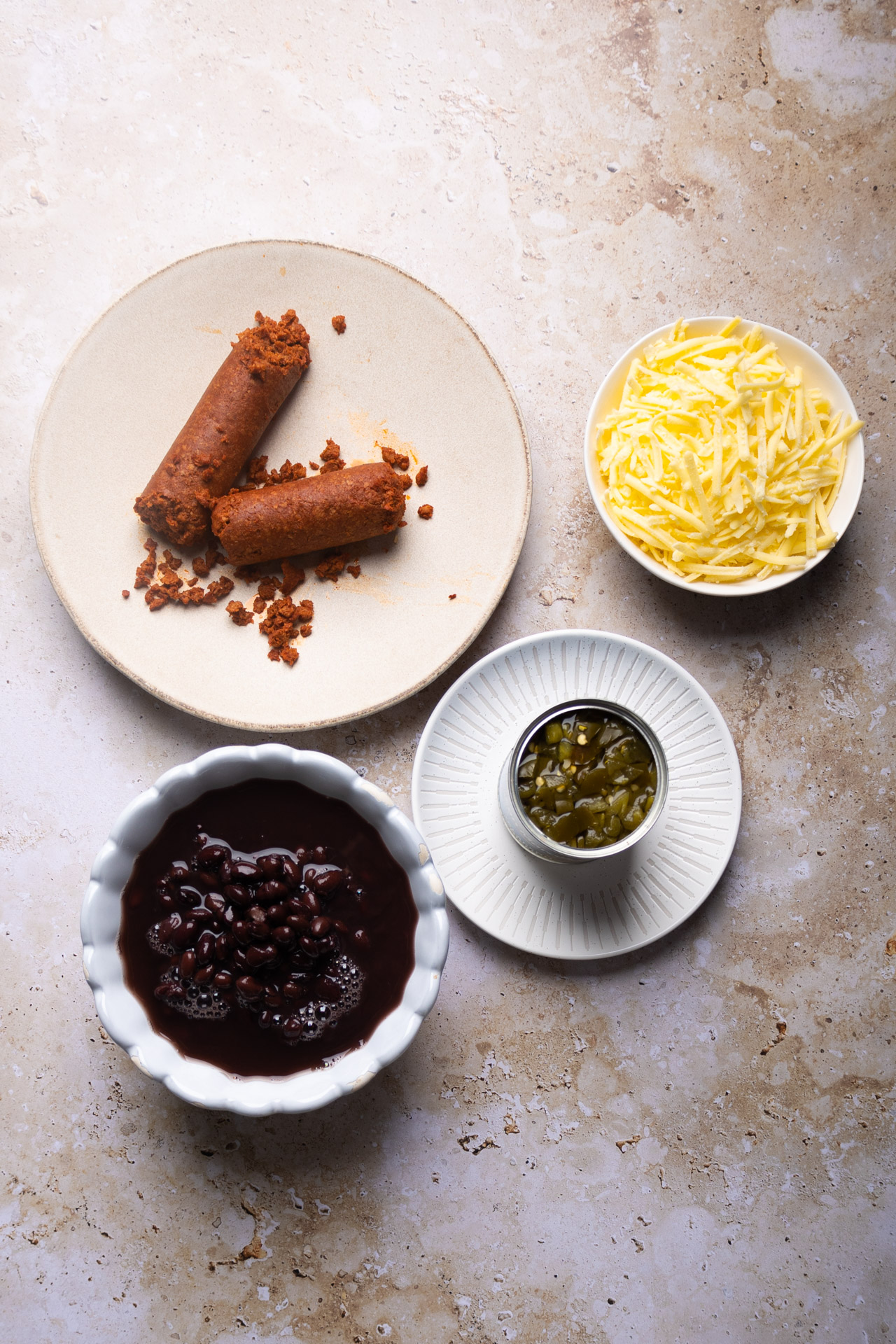 Tres platos y un cuenco sobre una superficie de color beige claro. En un plato hay chorizo cocido desmenuzado, en otro hay queso amarillo rallado, en un cuenco hay frijoles puercos y en un plato pequeño hay chiles verdes picados.