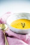 A textured white bowl filled with creamy roasted butternut squash soup (vegan), garnished with a sprig of sage and sprinkled with black pepper. The bowl rests on a soft pink cloth alongside a shiny gold spoon, all set against a bright, minimalist background.