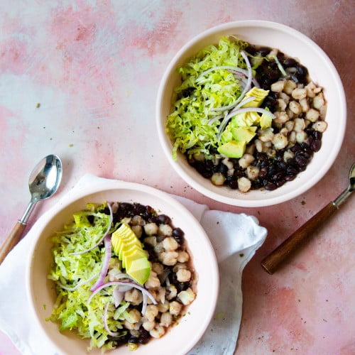 Dos tazones de sopa de frijoles negros y aguacate, también conocida como pozole con frijol negro, sobre fondo rosa.