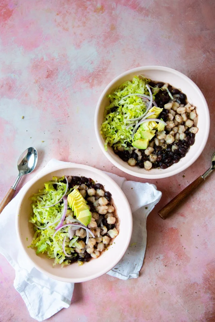 Dos tazones de sopa de frijoles negros y aguacate, también conocida como pozole con frijol negro, sobre fondo rosa.