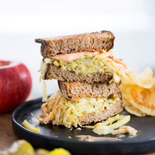 Un sándwich Reuben con pan de centeno, ensalada de col rallada y salsa se corta por la mitad y se sirve en un plato negro. Acompañado de patatas fritas crujientes y pepinillos, una manzana se encuentra al fondo, sobre una superficie de madera.