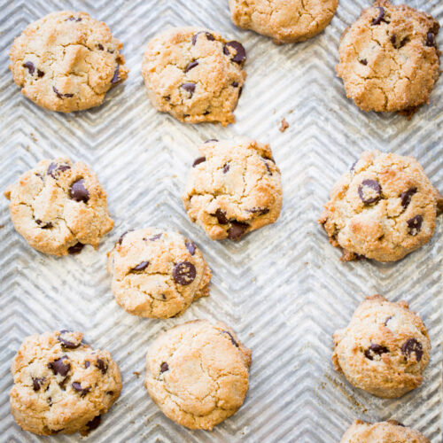 Vista aérea de varias galletas doradas con harina de almendras, distribuidas uniformemente en una bandeja para hornear con textura en zigzag. Las galletas tienen chispas de chocolate visibles, superficies agrietadas y la bandeja muestra señales de uso.