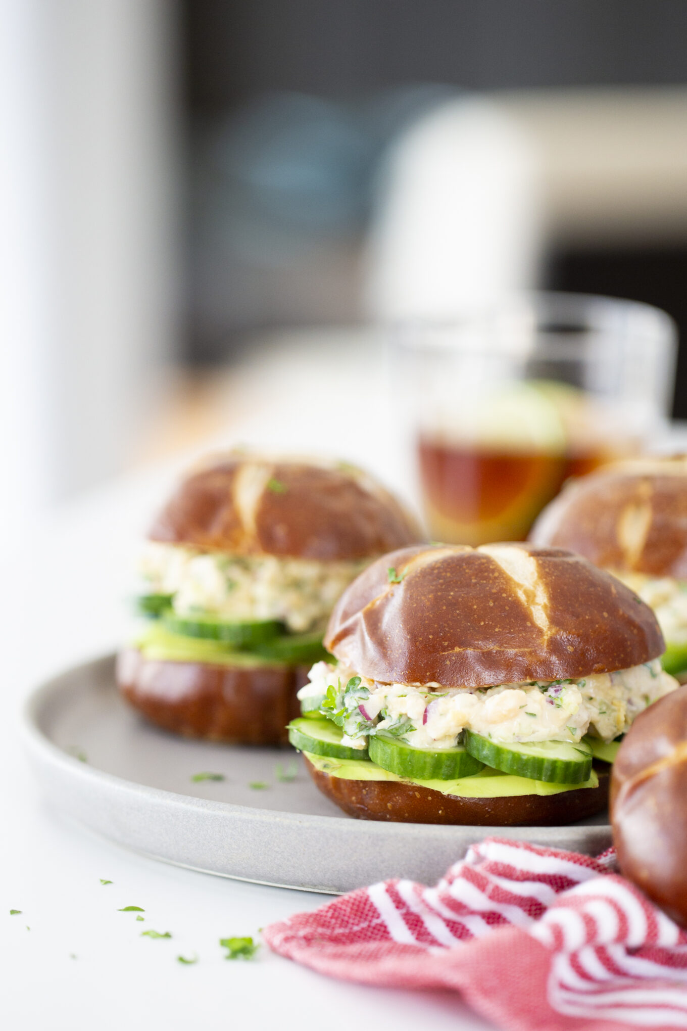 Tres sándwiches de atún veganos con pan de pretzel se sirven rellenos de una cremosa ensalada, rodajas de pepino y hierbas en un plato gris. Cerca, un vaso de té helado y una servilleta de tela de rayas rojas, sobre un fondo ligeramente difuminado.