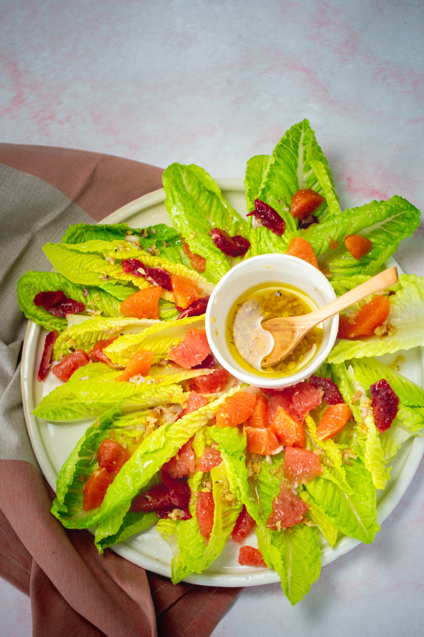 Un plato de hojas frescas de lechuga romana dispuestas en círculo con rodajas de pomelo y naranja sanguina crea una vibrante ensalada de cítricos. En el centro hay un pequeño tazón de vinagreta con una cuchara de madera, junto a una servilleta de tela rosa y beige.