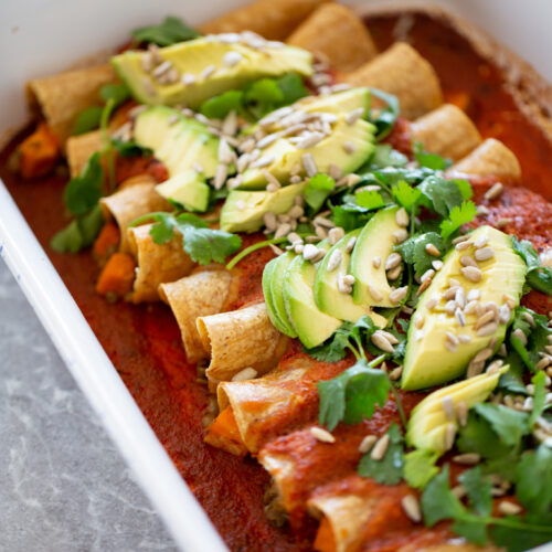 Un plato de enchiladas veganas cubiertas con rodajas de aguacate, cilantro y semillas servidas en una fuente de cerámica para hornear.