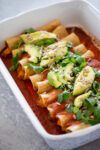 Un plato de enchiladas veganas cubiertas con rodajas de aguacate, cilantro y semillas, servidas en una fuente para horno blanca.