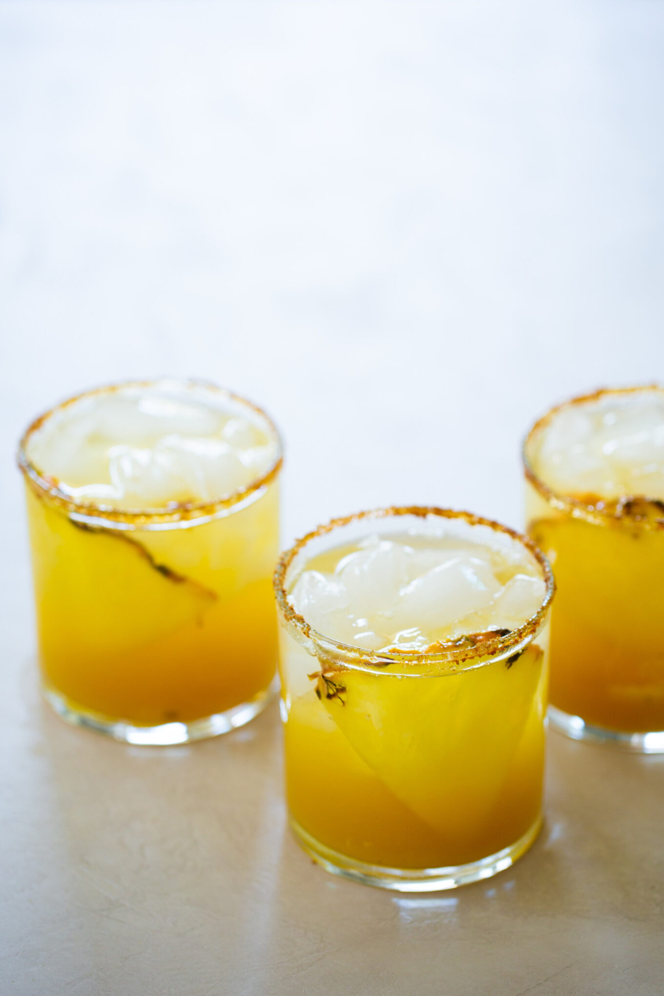 Tres vasos bajos, llenos de una bebida de color amarillo anaranjado brillante y hielo, se colocan sobre una superficie clara. Los bordes están cubiertos con una mezcla de especias, que recuerda a un cóctel sin alcohol de margarita de piña, adornado con una ramita de hierbas para un toque refrescante.