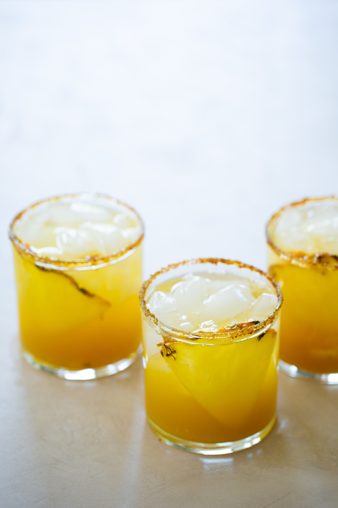 Tres vasos bajos, llenos de una bebida de color amarillo anaranjado brillante y hielo, se colocan sobre una superficie clara. Los bordes están cubiertos con una mezcla de especias, que recuerda a un cóctel sin alcohol de margarita de piña, adornado con una ramita de hierbas para un toque refrescante.