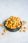 A small black bowl filled with chipotle roasted chickpeas sits on a light, textured surface. A few chickpeas are scattered around, displaying their golden-brown, crispy appearance and hinting at a savory, crunchy snack.
