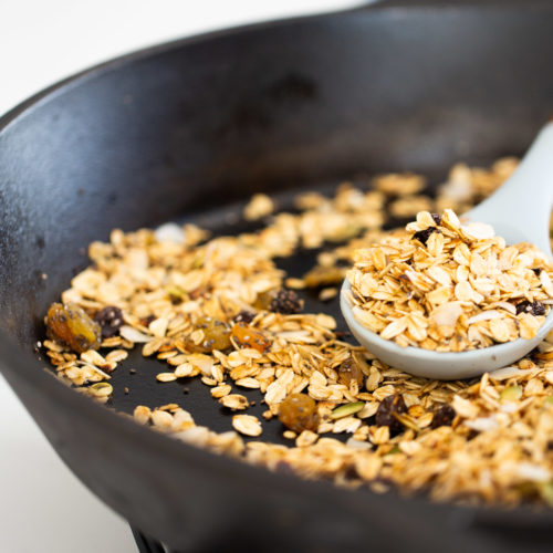 Primer plano de una sartén de hierro fundido negra sobre la estufa, con avena tostada mezclada con pasas. Una cuchara gris sirve una porción de la mezcla de avena. El fondo está desenfocado, resaltando el contenido de la sartén.