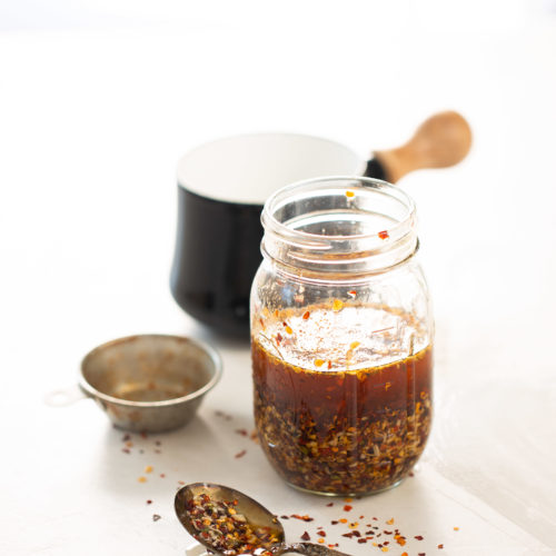 Sobre una superficie blanca hay un frasco de vidrio repleto de aceite picante de chile crujiente, acompañado de un colador de metal y una cuchara cargada de hojuelas de chile vibrantes. En el fondo suavemente difuminado, una taza negra con asa de madera realza esta atractiva escena de cocina centrada en delicias caseras.