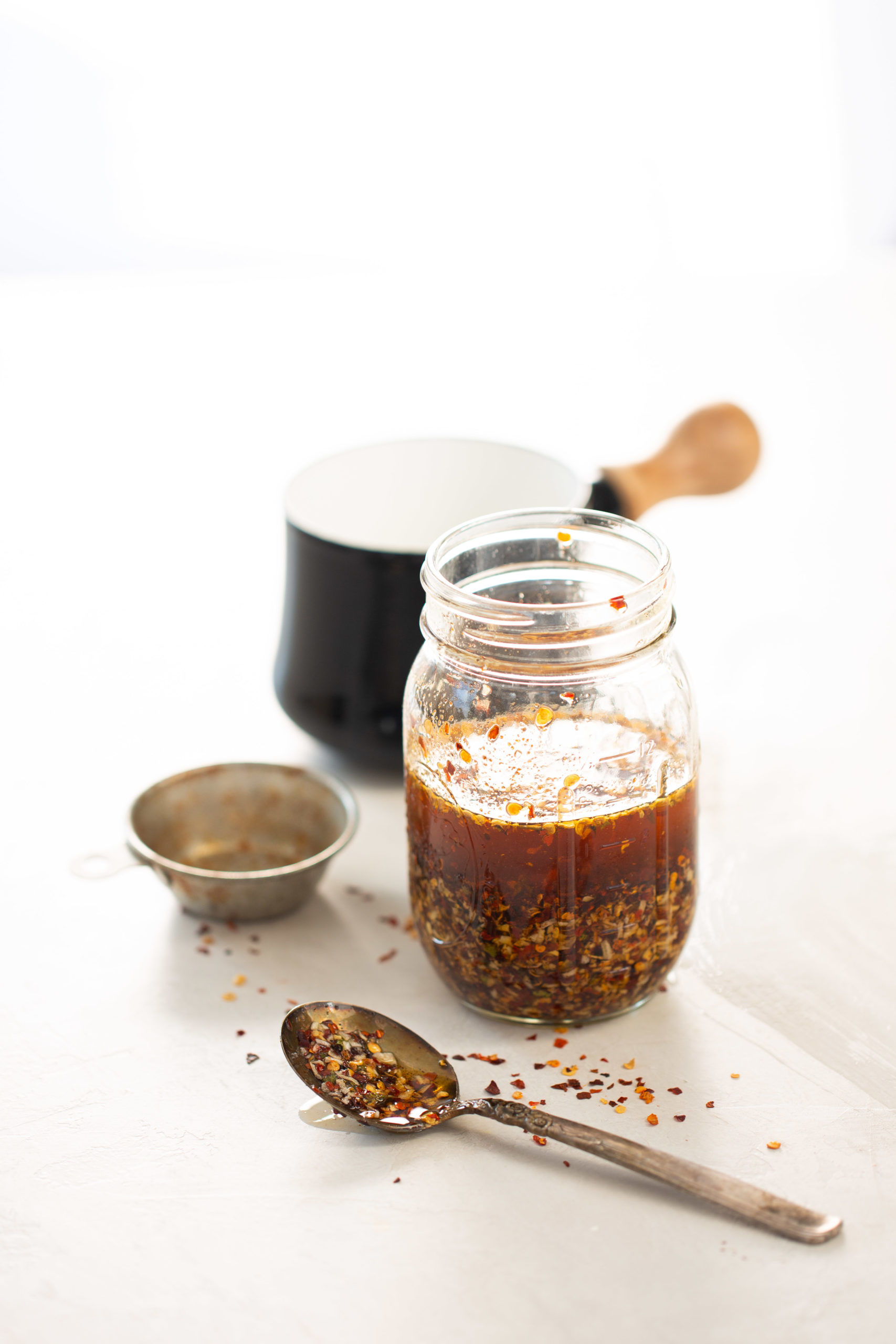 Sobre una superficie blanca hay un frasco de vidrio repleto de aceite picante de chile crujiente, acompañado de un colador de metal y una cuchara cargada de hojuelas de chile vibrantes. En el fondo suavemente difuminado, una taza negra con asa de madera realza esta atractiva escena de cocina centrada en delicias caseras.