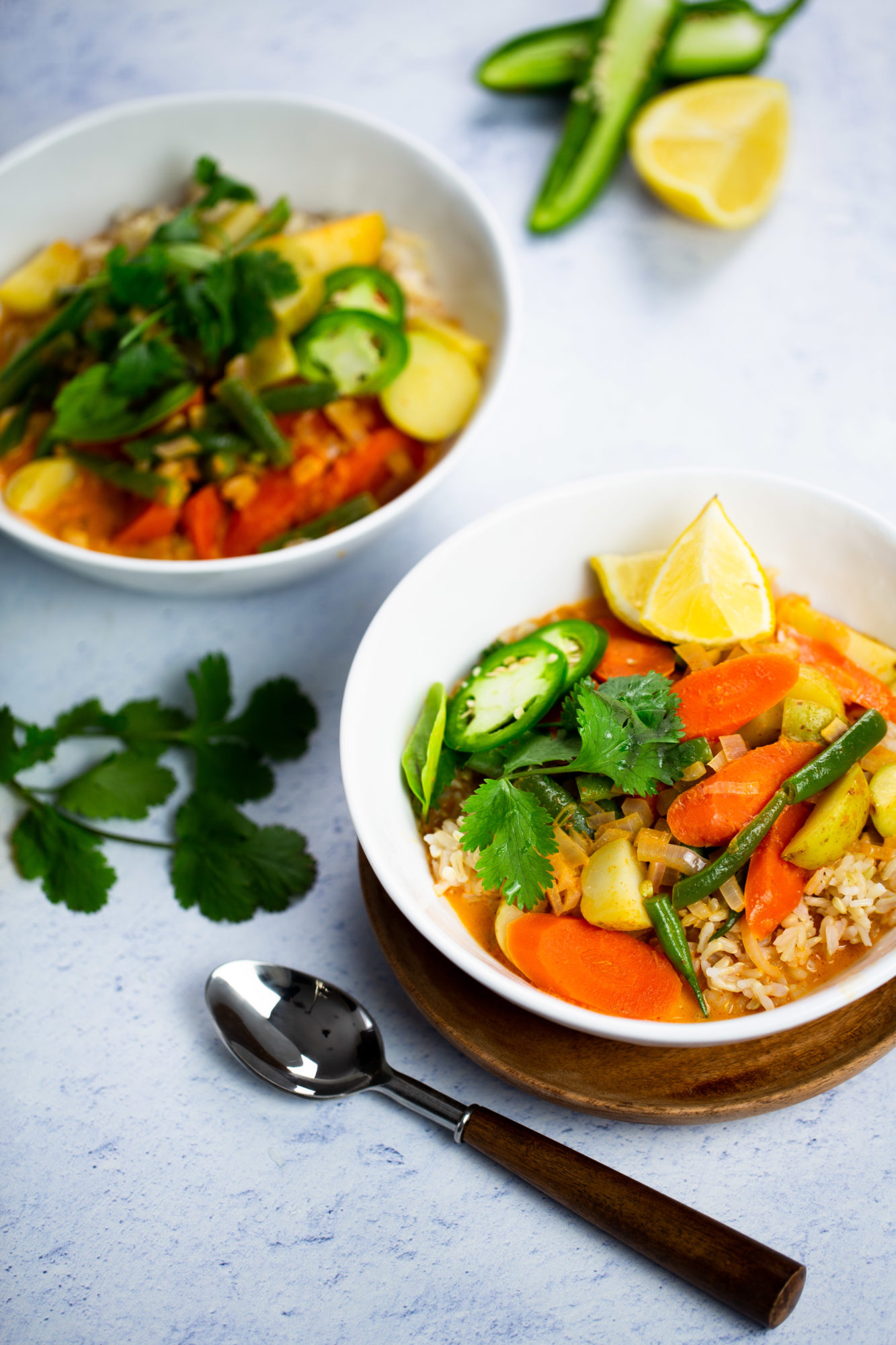 Dos tazones de curry de verduras tailandés con arroz se adornan con cilantro y jalapeños en rodajas, acompañados de gajos de lima. Estos vibrantes platos llevan zanahorias, calabacín y judías verdes. Una cuchara reposa junto a los tazones sobre una superficie blanca con textura, con ramitas de cilantro cerca para un toque de frescura rapidísima.