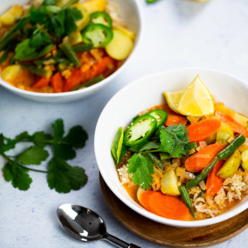 Dos tazones de curry de verduras tailandés con arroz se adornan con cilantro y jalapeños en rodajas, acompañados de gajos de lima. Estos vibrantes platos llevan zanahorias, calabacín y judías verdes. Una cuchara reposa junto a los tazones sobre una superficie blanca con textura, con ramitas de cilantro cerca para un toque de frescura rapidísima.