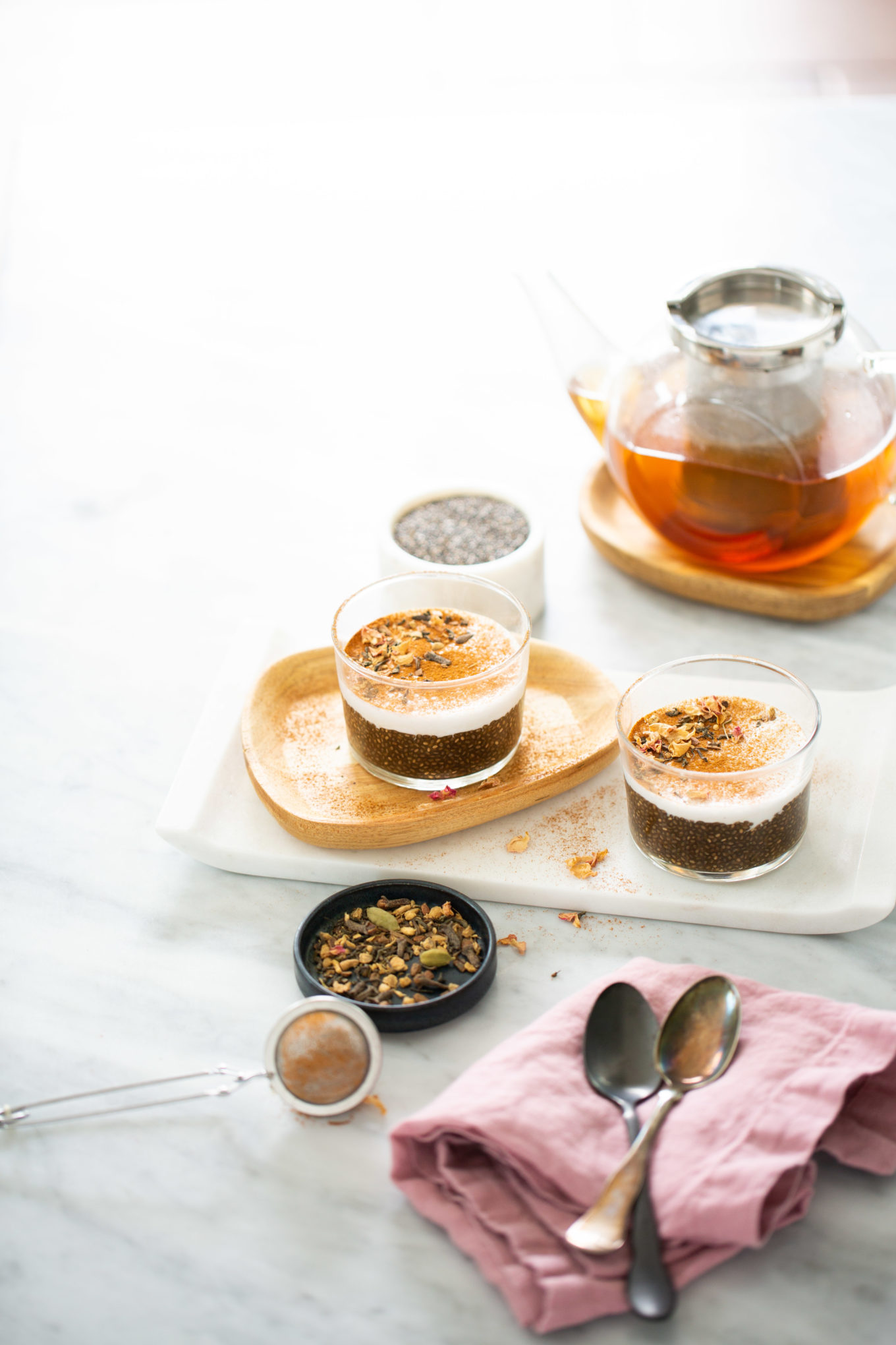 Dos vasos de pudín de chía infusionado con esencia de té chai y cubiertos con nueces descansan sobre una bandeja blanca. Cerca, un pequeño cuenco con semillas y un colador acompañan el decorado. Al fondo, una tetera de vidrio llena de té aromático reposa sobre un posavasos de madera. Dos cucharas descansan sobre una servilleta rosa doblada en primer plano sobre la superficie de mármol blanco.