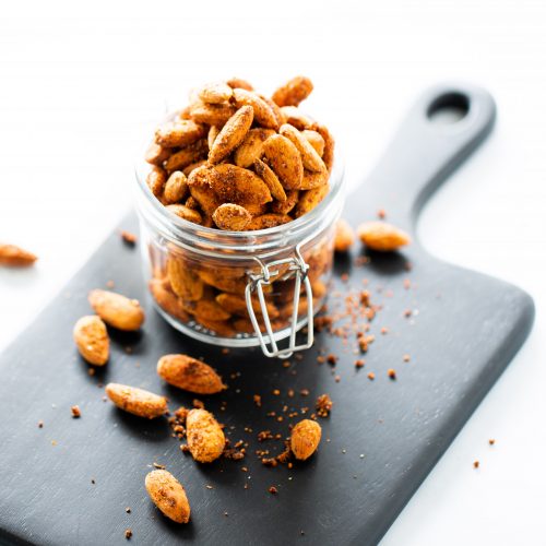 Un frasco de vidrio lleno de almendras veganas tostadas y picantes reposa sobre una tabla de cortar negra. Hay algunas almendras y migas esparcidas alrededor del frasco. El fondo es blanco y está bien iluminado, lo que le da un aspecto limpio y minimalista.