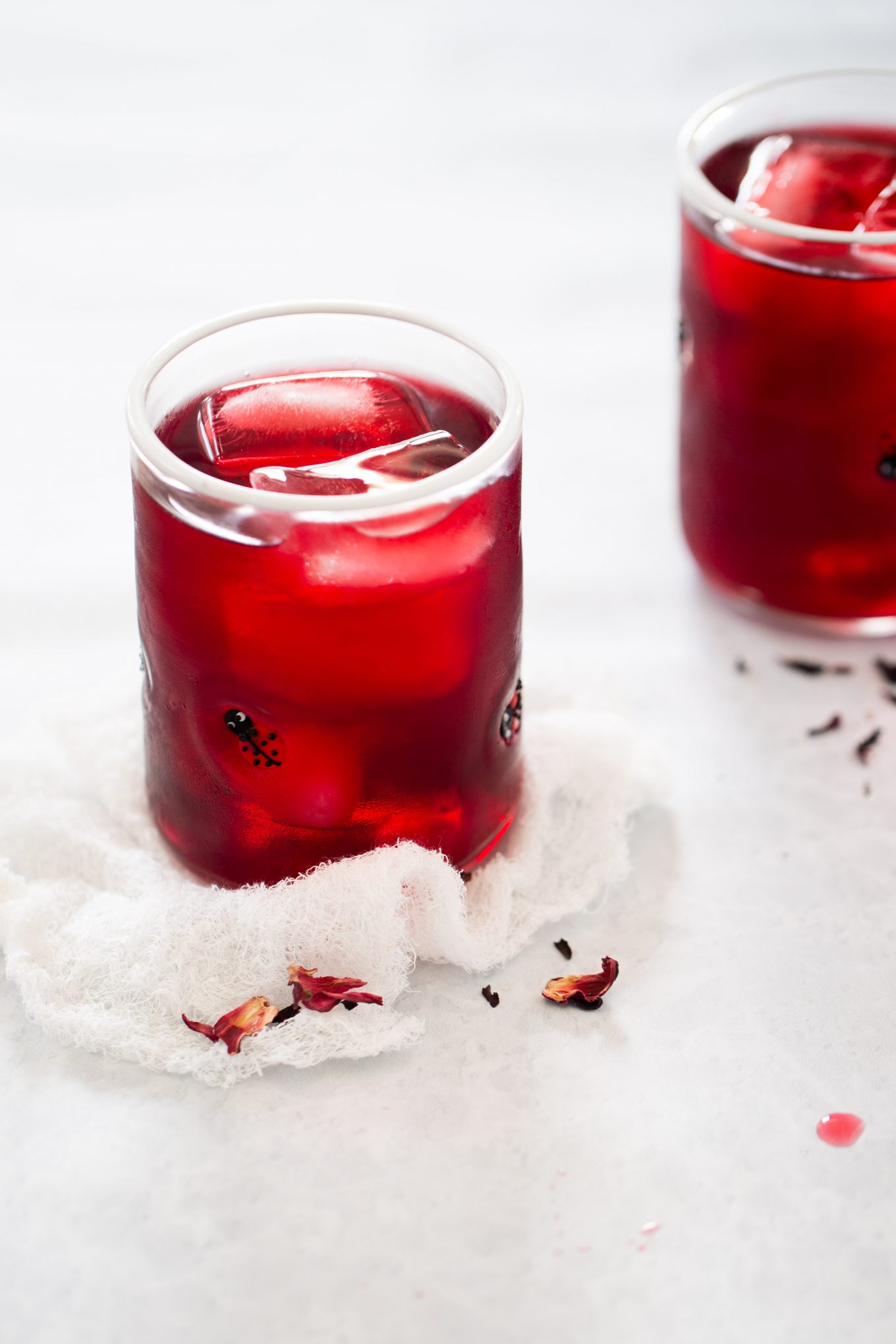 Primer plano de un vaso lleno de una bebida roja y cubitos de hielo, colocado sobre una tela de color claro. Otro vaso similar está ligeramente desenfocado en el fondo. Hay pétalos de hibisco secos esparcidos alrededor de los vasos, lo que sugiere una bebida de agua de Jamaica. El fondo es una superficie gris clara.