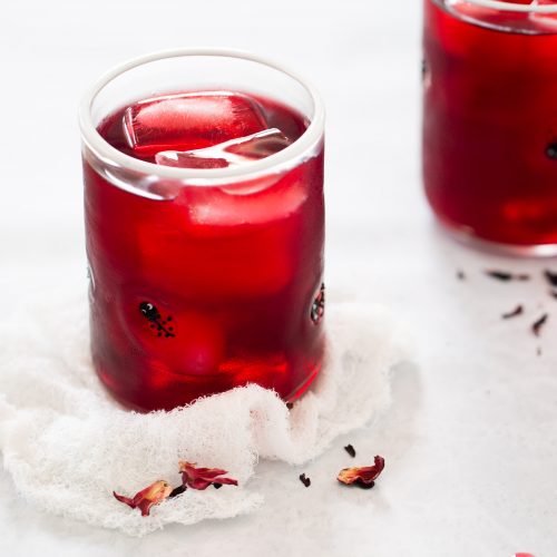 Primer plano de un vaso lleno de una bebida roja y cubitos de hielo, colocado sobre una tela de color claro. Otro vaso similar está ligeramente desenfocado en el fondo. Hay pétalos de hibisco secos esparcidos alrededor de los vasos, lo que sugiere una bebida de agua de Jamaica. El fondo es una superficie gris clara.