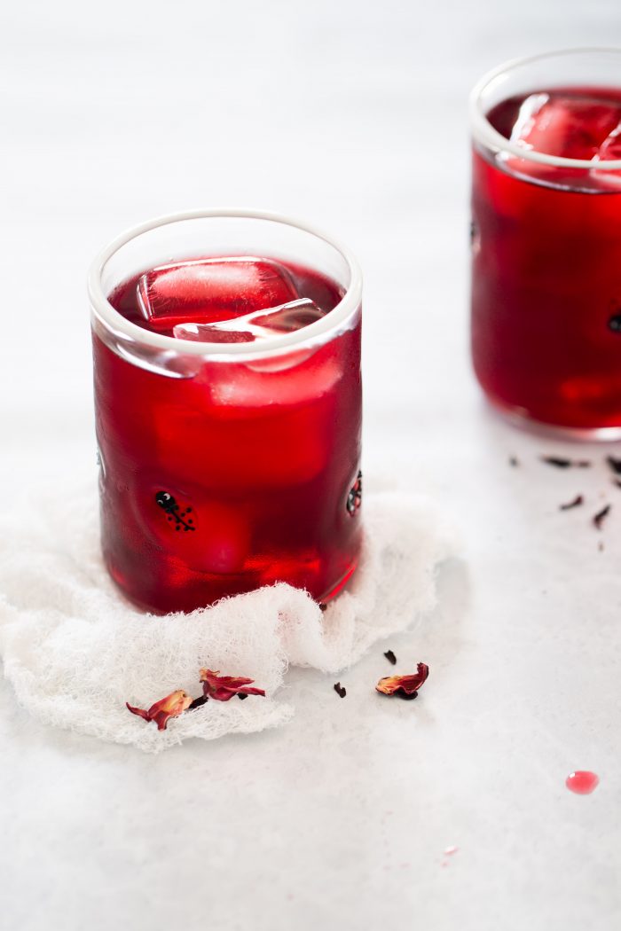 Primer plano de un vaso lleno de una bebida roja y cubitos de hielo, colocado sobre una tela de color claro. Otro vaso similar est&aacute; ligeramente desenfocado en el fondo. Hay p&eacute;talos de hibisco secos esparcidos alrededor de los vasos, lo que sugiere una bebida de agua de Jamaica. El fondo es una superficie gris clara.