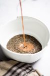Un cuenco blanco repleto de semillas de chía se rocía con leche chocolatada, formando el comienzo de un delicioso pudín de chocolate y chía. El líquido, a mitad de movimiento, salpica suavemente las semillas. Un paño rayado se asoma a la vista sobre la superficie blanca prístina.