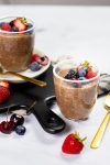 Dos vasos de rico pudín de chocolate y chía están adornados con bayas frescas, como fresas, arándanos y frambuesas, sobre una superficie blanca. Cerca hay una cuchara dorada. Hay más bayas esparcidas por todas partes y una tabla de servir negra es parcialmente visible en el fondo.