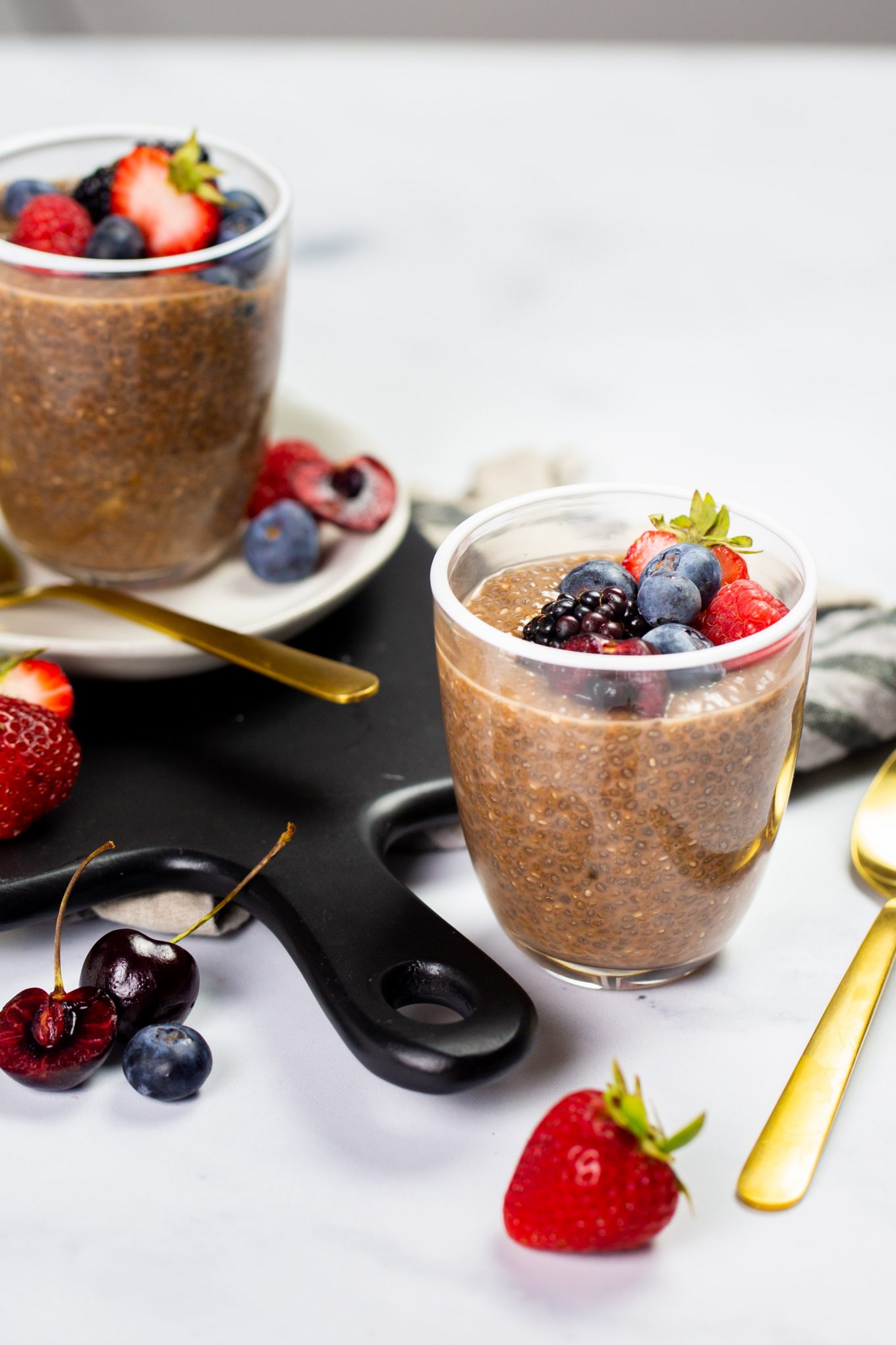 Dos vasos de rico pudín de chocolate y chía están adornados con bayas frescas, como fresas, arándanos y frambuesas, sobre una superficie blanca. Cerca hay una cuchara dorada. Hay más bayas esparcidas por todas partes y una tabla de servir negra es parcialmente visible en el fondo.