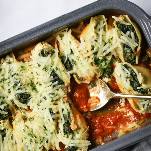 Una fuente para hornear que contiene conchas de pasta horneadas con espinacas y queso encima, visiblemente adornadas con hierbas. Hay una cuchara para servir dentro del plato, que reposa sobre una superficie blanca con un paño gris.