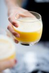 Close-up of a hand holding a round glass filled with creamy, frothy pineapple ginger juice, its vibrant orange-yellow hue inviting. The drink boasts a smooth, foamy top. Another blurred glass is visible in the foreground, suggesting a lively social setting against the soft-focus background.