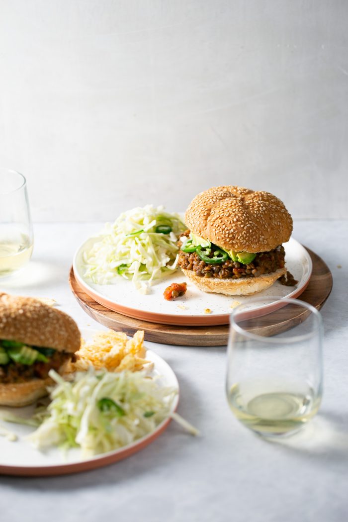 Dos platos contienen cada uno un pan de hamburguesa con semillas de sésamo relleno de una hamburguesa al estilo Sloppy Joes, lechuga y rodajas de aguacate, servido con col rallada. Dos copas de vino blanco reposan cerca, sobre una superficie clara con un fondo neutro.