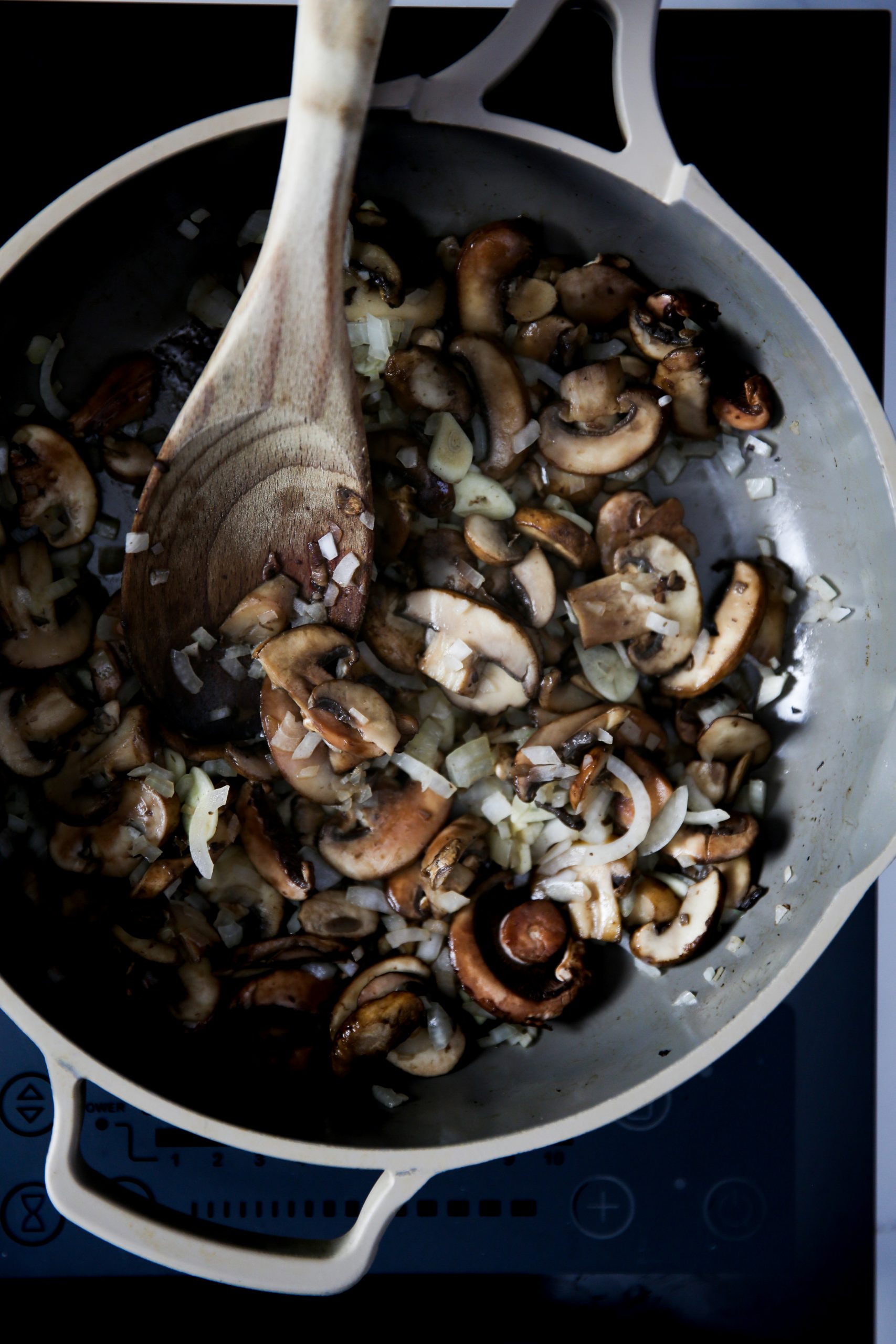 Una sartén blanca sobre el fuego está llena de champiñones y cebollas picados que se saltean para los tacos gobernador veganos. Una cuchara de madera se apoya en la sartén, revolviendo la mezcla, mientras que la iluminación superior destaca las vibrantes verduras y la sartén.