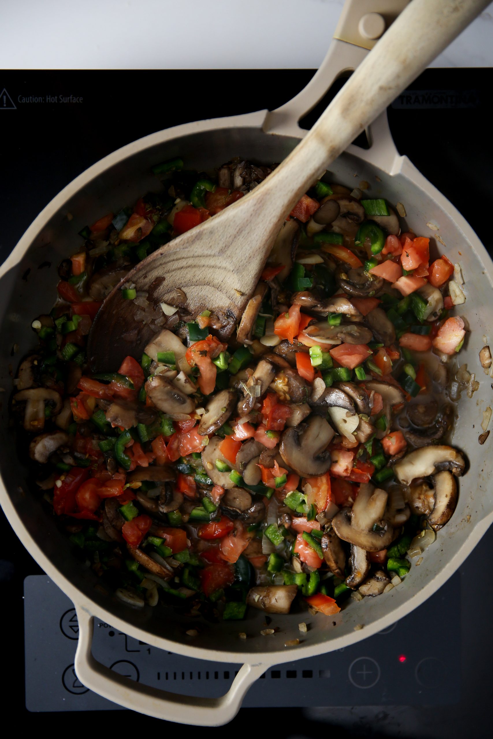 Una sartén beige sobre la estufa contiene champiñones salteados, tomates cortados en dados y cebollas verdes en rodajas para tacos gobernador veganos, que se remueven con una cuchara de madera. Las coloridas verduras parecen recién cocinadas. Los mandos de la cocina y la etiqueta "Precaución: Superficie caliente".