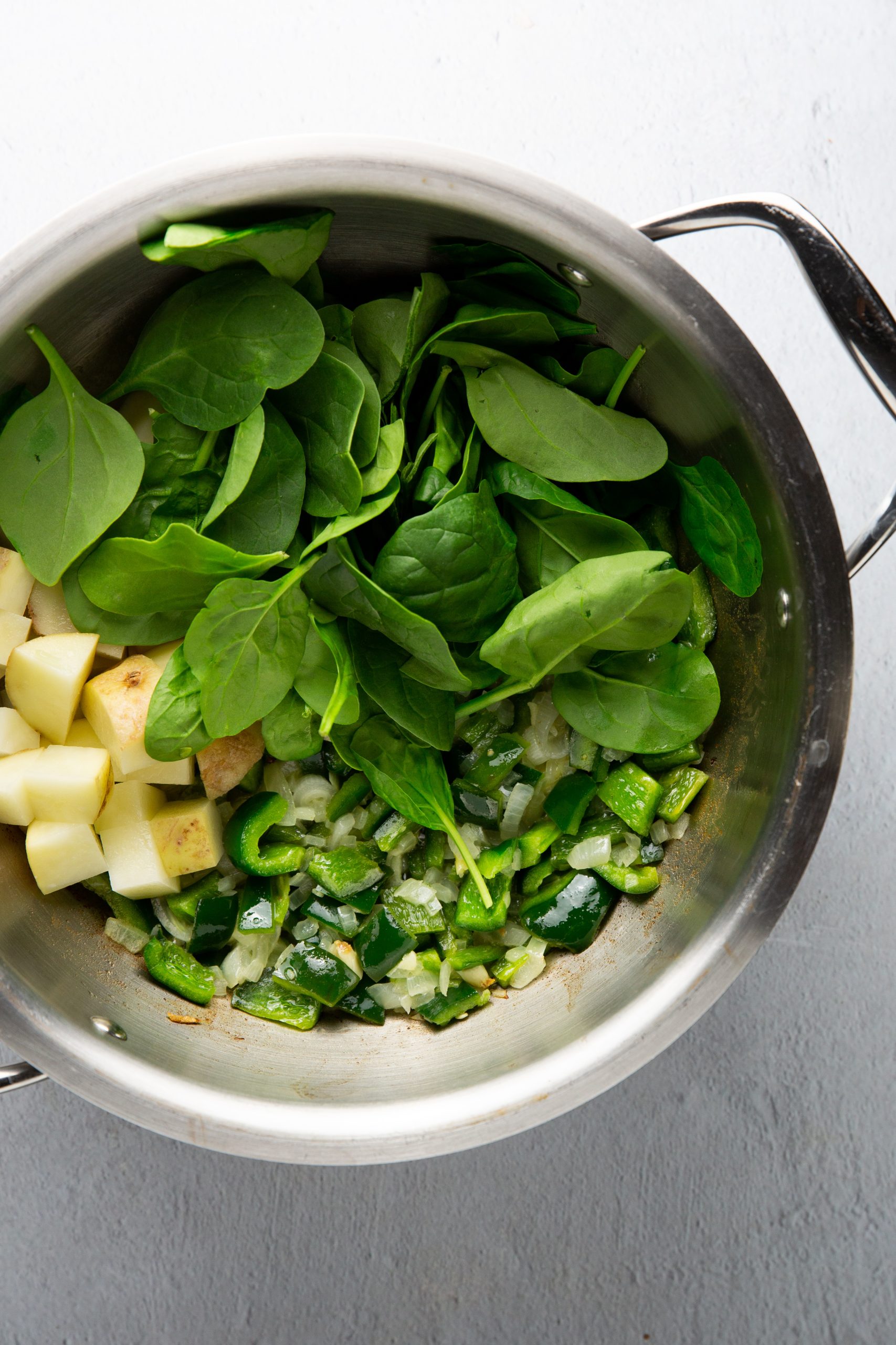 Una olla de acero inoxidable contiene hojas de espinacas frescas, patatas cortadas en dados, poblano y espinacas picados, pimiento verde y cebollas salteadas, todo dispuesto en secciones separadas. Las verduras aparecen crudas y están listas para su posterior cocción.