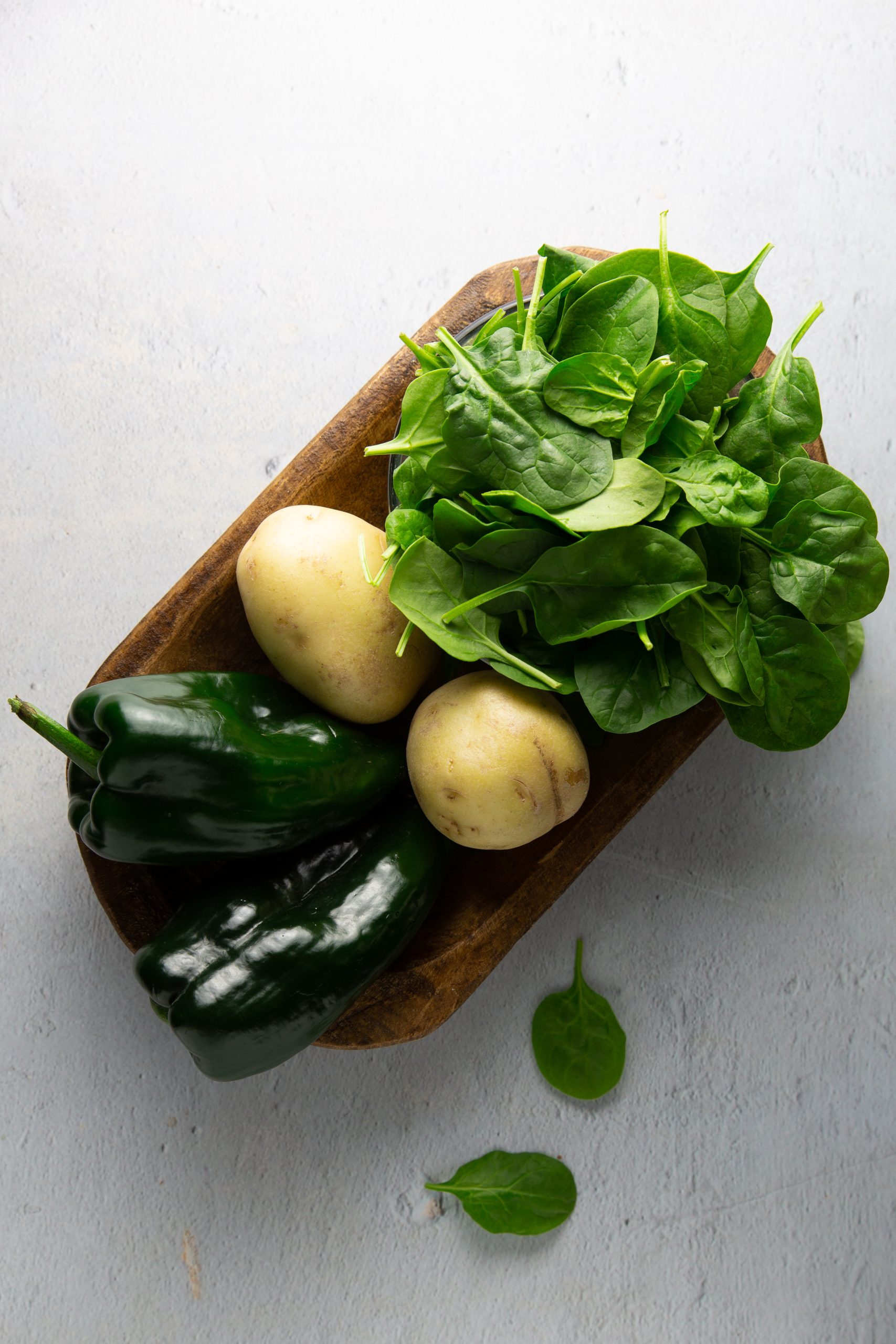 Una bandeja de madera contiene hojas frescas de espinaca, dos papas amarillo pálido y dos chiles poblanos verde oscuro, perfectos para un plato de poblano y espinacas. Unas cuantas hojas de espinaca están esparcidas por la superficie de textura gris clara junto a la bandeja.