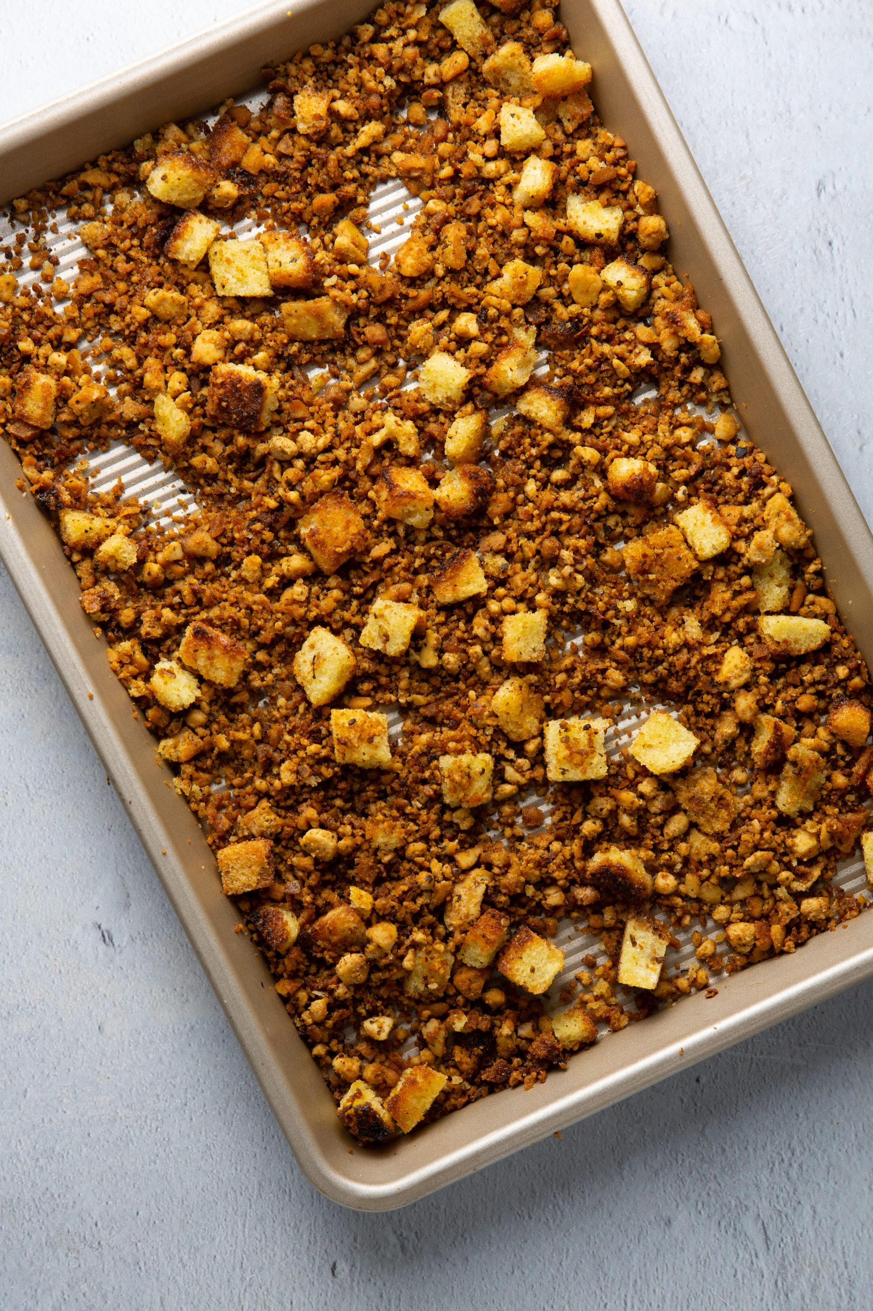 Una bandeja para hornear sostiene una capa uniforme de mezcla de relleno dorado con trocitos de pan, poblano y espinacas, tostados y sazonados. La bandeja descansa sobre una superficie gris claro, y el relleno aparece crujiente y bien horneado.