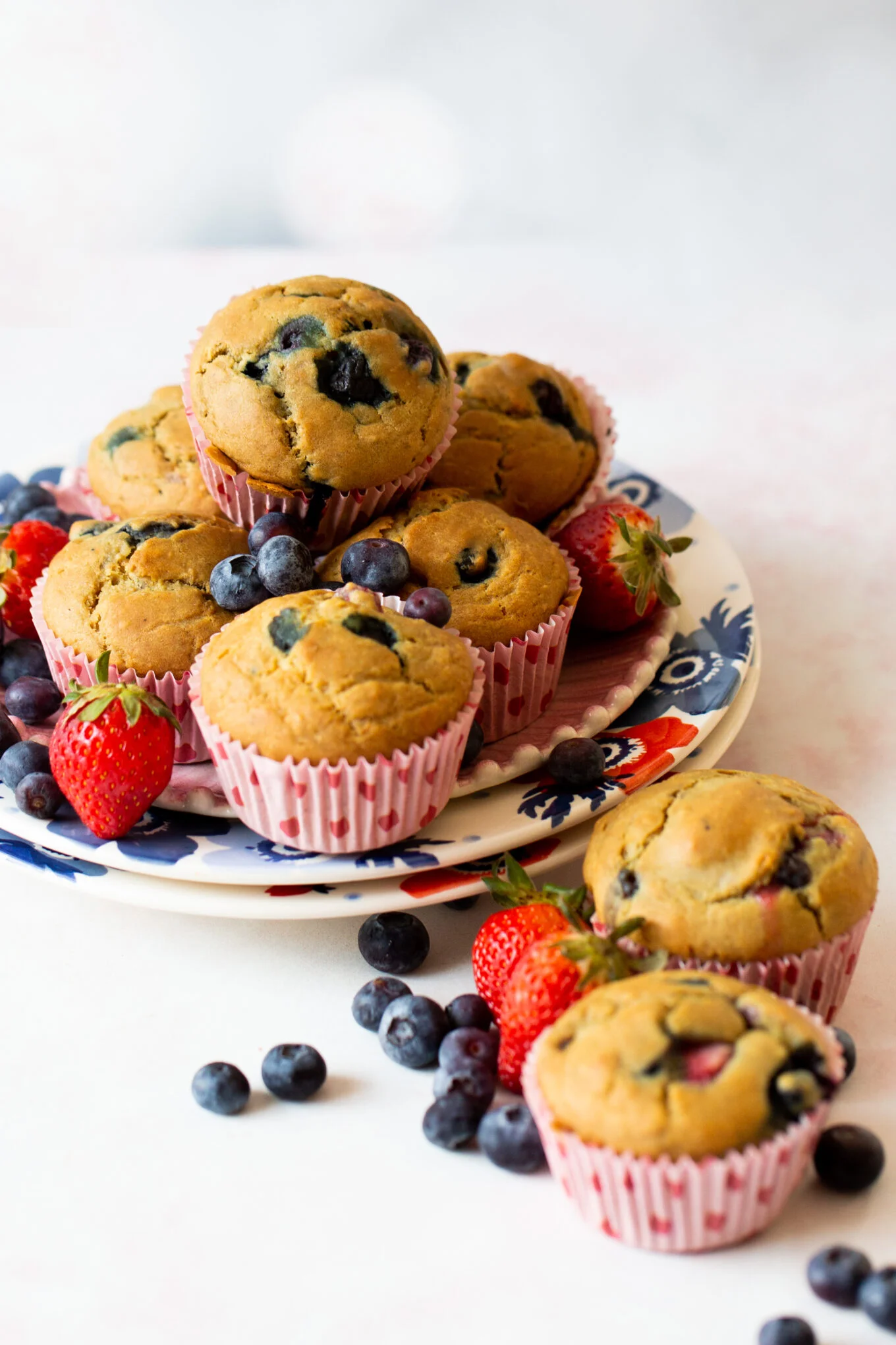 Un plato de muffins de frutos rojos recién horneados sobre una vajilla decorativa. Los muffins, en moldes de rayas rosas, rebosan de arándanos. A su alrededor, frutos rojos frescos como arándanos y fresas. La escena se desarrolla sobre un fondo suave y claro, creando un cuadro encantador.